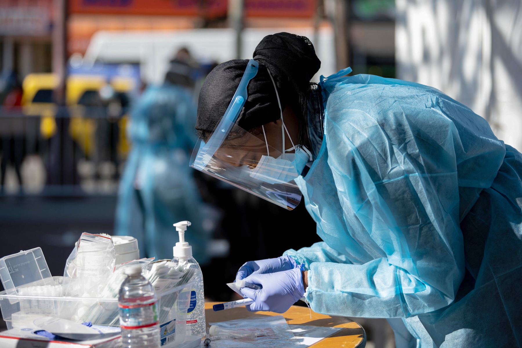 A health worker in a face shield and gown labels a test sample at an outdoor testing table